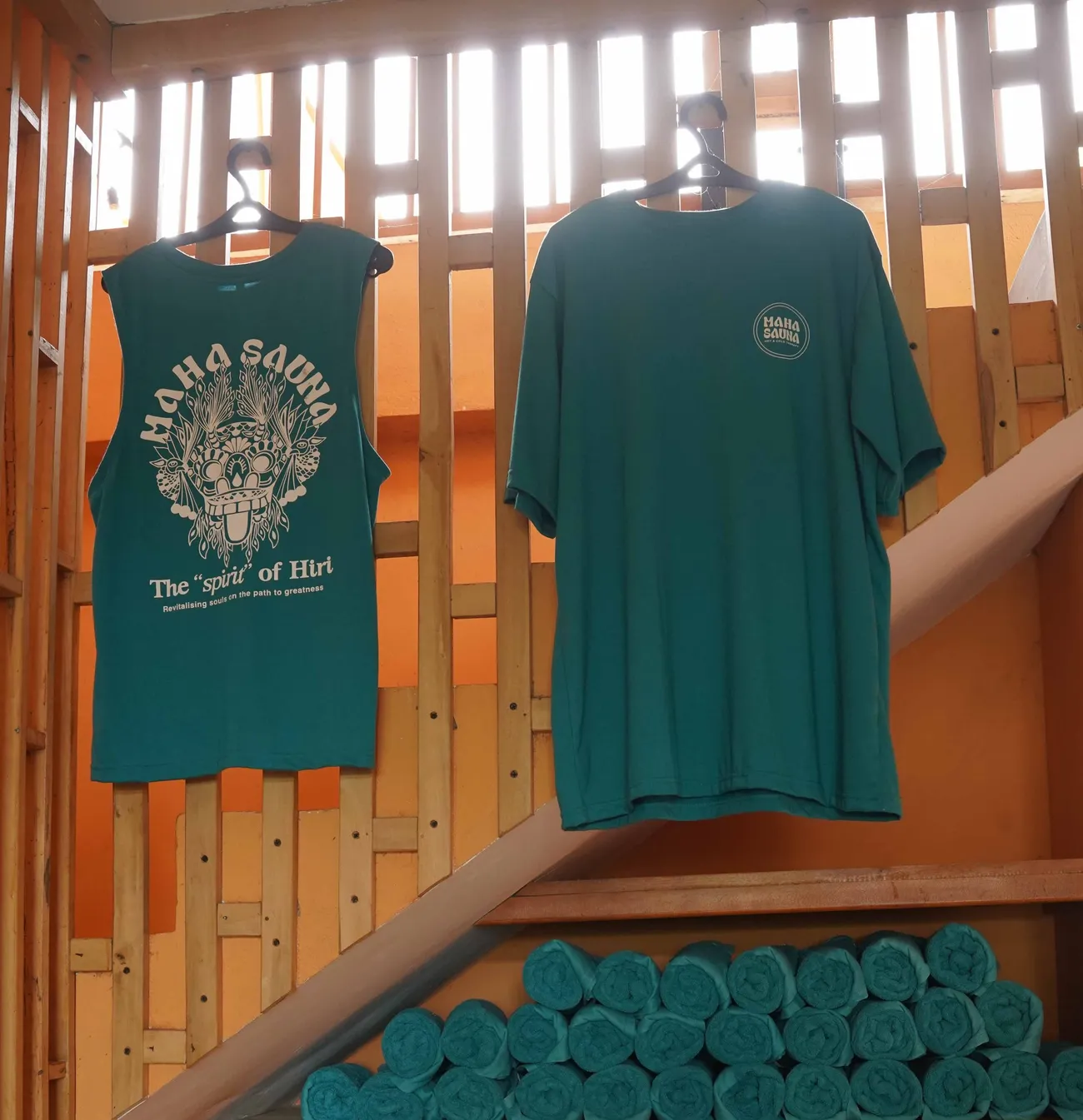 The Spirit of Hiri tank and the Maha Sauna tee hanging above a shelf of rolled towels